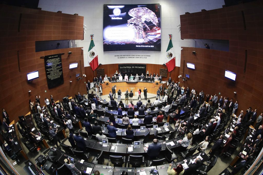 La imagen muestra una sesión del Congreso de México con legisladores y asistentes en un auditorio grande, con banderas mexicanas y pantallas que muestran información relevante.