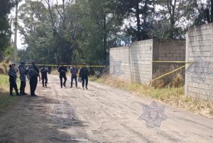 Persona en la escena de una investigación policial en una calle de tierra, con varios oficiales y una cerca de bloques de cemento, en un ambiente de investigación y seguridad pública.
