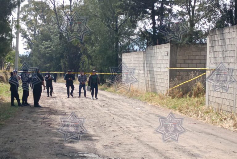 Persona en la escena de una investigación policial en una calle de tierra, con varios oficiales y una cerca de bloques de cemento, en un ambiente de investigación y seguridad pública.