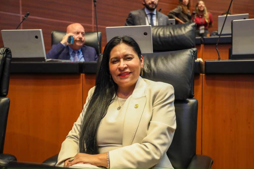 Mujer en una reunión formal en un entorno de oficina con otros asistentes en el fondo, vestida con traje beige y sonriendo.