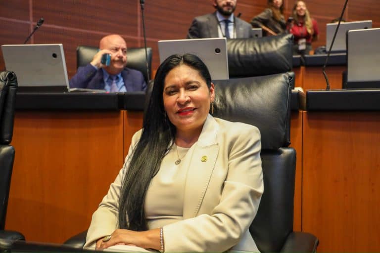 Mujer en una reunión formal en un entorno de oficina con otros asistentes en el fondo, vestida con traje beige y sonriendo.