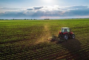 Cultivador agrícola trabajando en un campo de cultivo verde bajo un cielo nublado con rayos de sol.