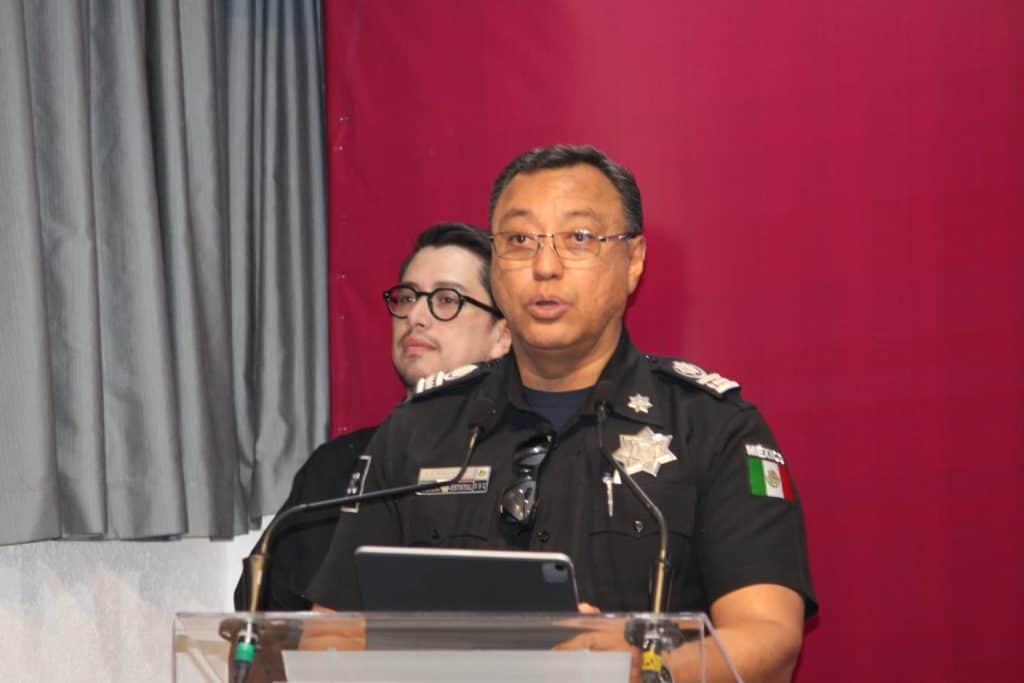 Policía mexicana en uniforme durante una conferencia de prensa, con fondo rojo y cortinas grises a un lado.