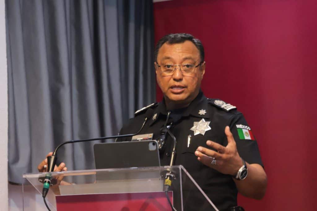 Oficial de policía hablando en una conferencia, vestido con uniforme y en un escenario con cortinas grises y fondo rojo.