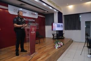 Policía en uniforme dando un discurso en un evento oficial en un auditorio con pantalla y decoraciones florales.