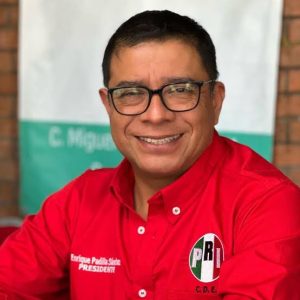 Candidato en campaña con camiseta roja y gafas, sonriendo, en un entorno exterior con fondo de pared y cartel.