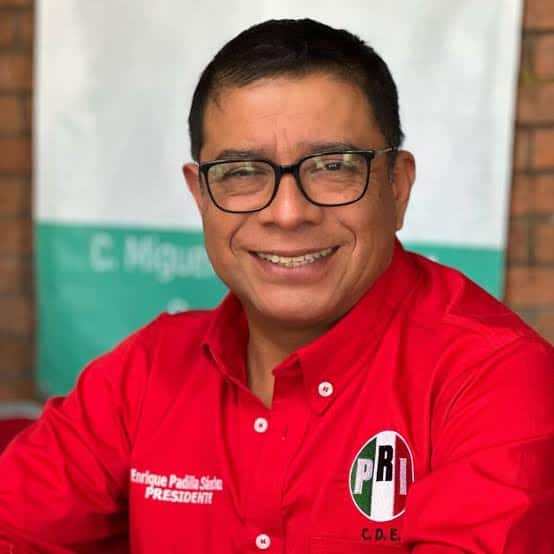 Candidato en campaña con camiseta roja y gafas, sonriendo, en un entorno exterior con fondo de pared y cartel.