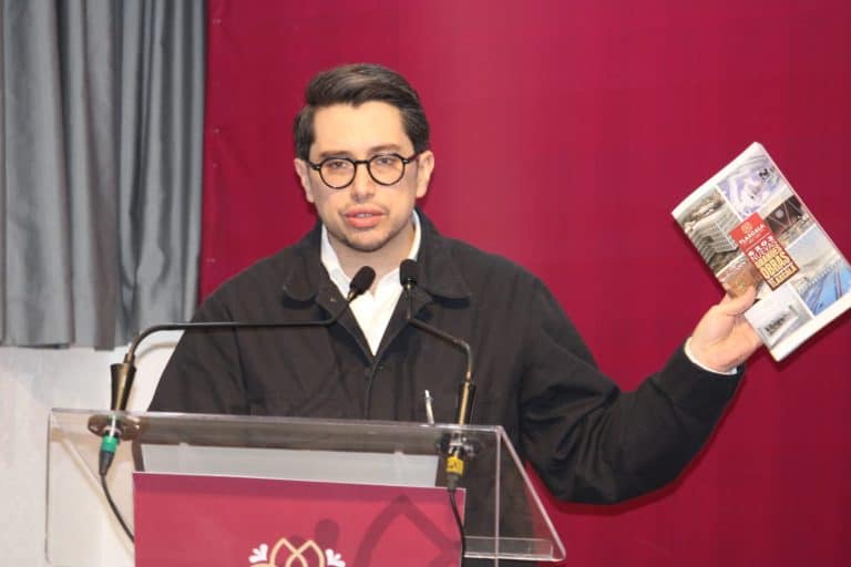 Hombre con gafas y chaqueta negra, hablando en un podio con fondo rojo y sosteniendo un periódico en la mano derecha.