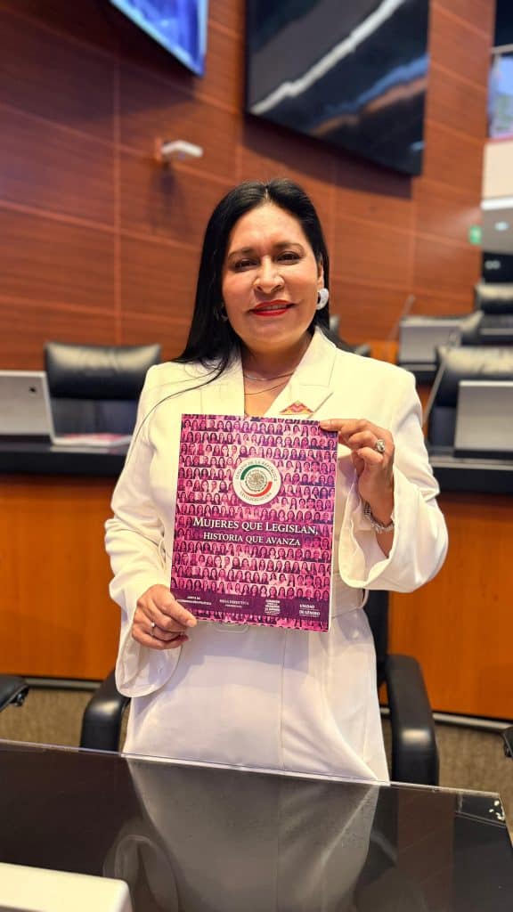 Mujer en una sala de reuniones sosteniendo un libro con portada rosa y texto en español, vestida con traje blanco y cabello largo y oscuro.