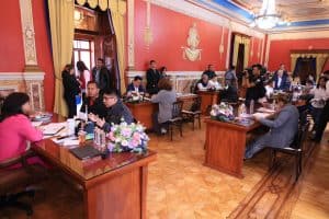 Reunión formal en una sala elegante con varias personas sentadas en mesas, discutiendo y tomando notas durante una conferencia o evento oficial en un ambiente decorado con detalles clásicos y ornament.