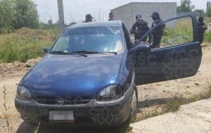 Un vehículo azul con la puerta del conductor abierta, rodeado por oficiales de seguridad en un área rural, posiblemente en una escena de investigación o intervención policial en Tlaxcala, México.