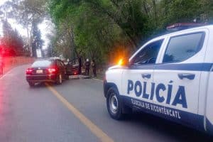 Un vehículo de policía de Tlaxcala de Xicohténcatl atiende un accidente en una carretera rural rodeada de árboles, con varias personas en la escena y un auto detenido en medio de la vía.