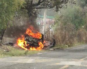 Vehículo incendiado a un lado de la carretera en Tlaxcala, con llamas y humo visible, junto a áreas verdes y árboles.