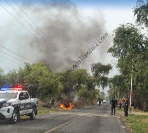 Línea 1: Vehículo en llamas en la carretera, con humo denso en el cielo y presencia de policías en la escena.