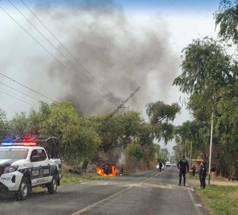 Línea 1: Vehículo en llamas en la carretera, con humo denso en el cielo y presencia de policías en la escena.