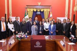 La imagen muestra a un grupo de funcionarios y representantes en una reunión oficial en un salón decorado con banderas y un escudo de Tlaxcala, en un evento formal.