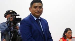 Persona en traje formal azul, en un evento público, con un fondo blanco y una cámara de video en el lado izquierdo.