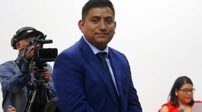 Persona en traje formal azul, en un evento público, con un fondo blanco y una cámara de video en el lado izquierdo.