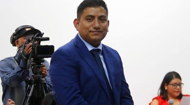 Persona en traje formal azul, en un evento público, con un fondo blanco y una cámara de video en el lado izquierdo.