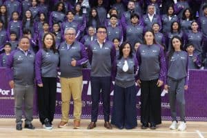 Una grupo de personas, incluyendo adultos y jóvenes, posando en un evento deportivo con uniformes morados y grises, en un gimnasio o cancha cubierta.