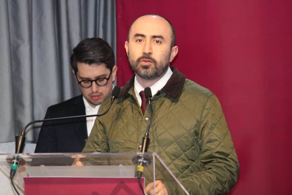 Hombre con barba y saco verde hablando en un podio durante una conferencia o evento formal.
