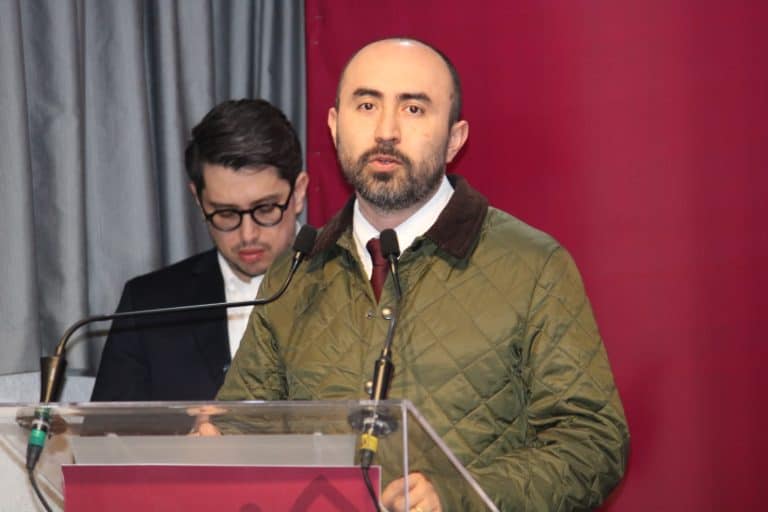 Hombre con barba y saco verde hablando en un podio durante una conferencia o evento formal.
