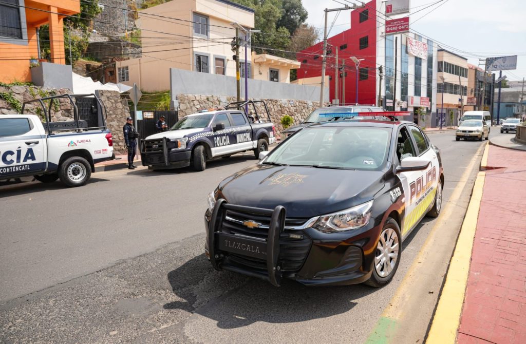 Vehículos de policía en una calle de Tlaxcala, México, con varias unidades estacionadas y un edificio moderno al fondo, durante el día.