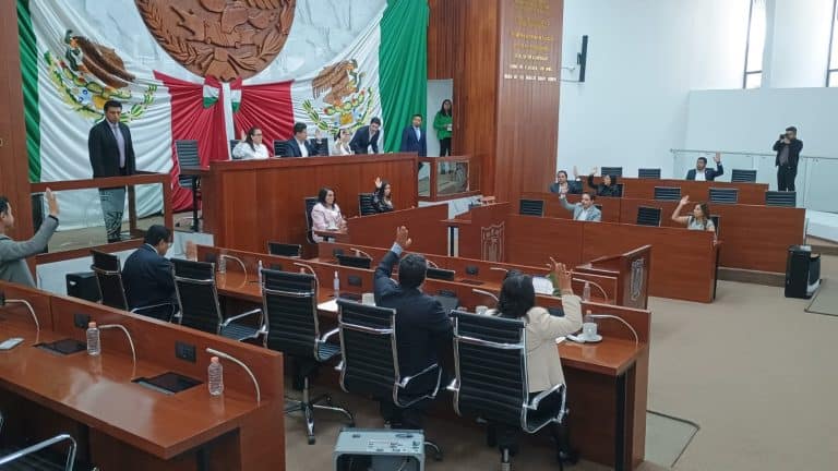 Sesión en el Congreso de Tlaxcala con legisladores y un gran escudo de México en el fondo, en un ambiente formal y de discusión política.