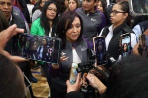 Una mujer en el centro, rodeada de periodistas y cámaras, da una declaración en un evento público, mientras varias personas la toman fotos y videos con sus teléfonos móviles.