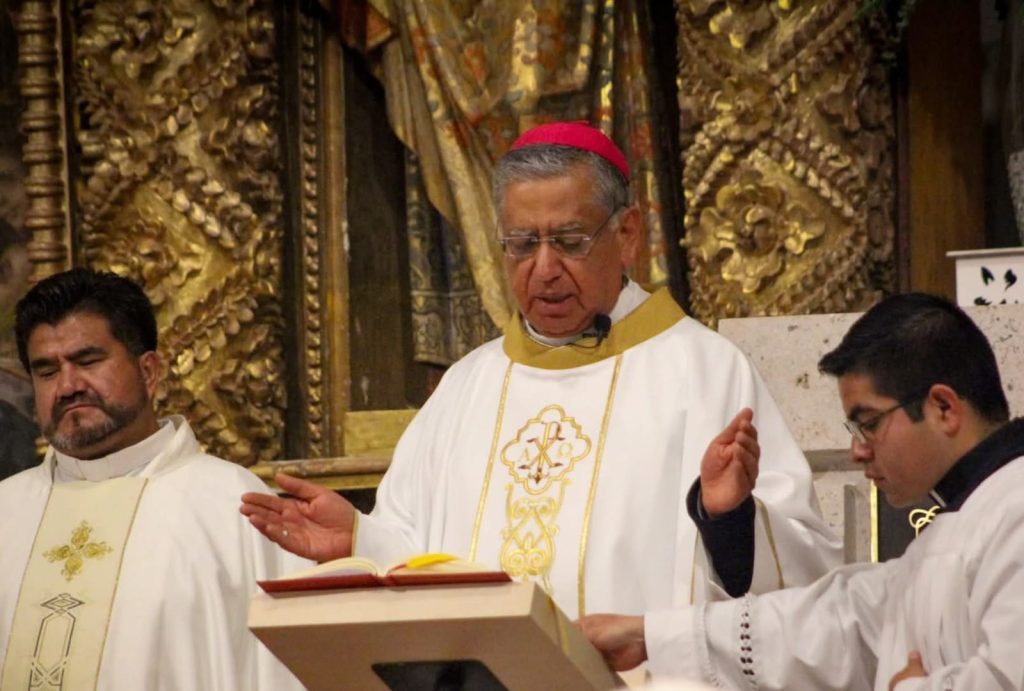 Obispo católico en ceremonia religiosa, vestido con vestimentas tradicionales y rodeado de otros sacerdotes en un entorno decorado con detalles dorados y religiosos.