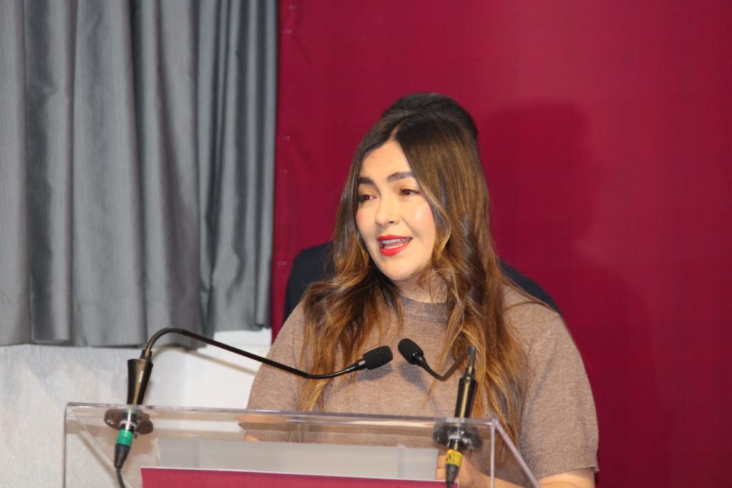 Amplia mujer con cabello castaño claro y ondulado, vestida con suéter beige, hablando en un podio con micrófonos en un evento formal, con fondo de cortinas grises y pared rosa oscuro.