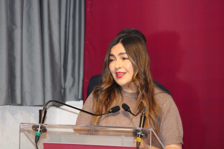 Amplia mujer con cabello castaño claro y ondulado, vestida con suéter beige, hablando en un podio con micrófonos en un evento formal, con fondo de cortinas grises y pared rosa oscuro.