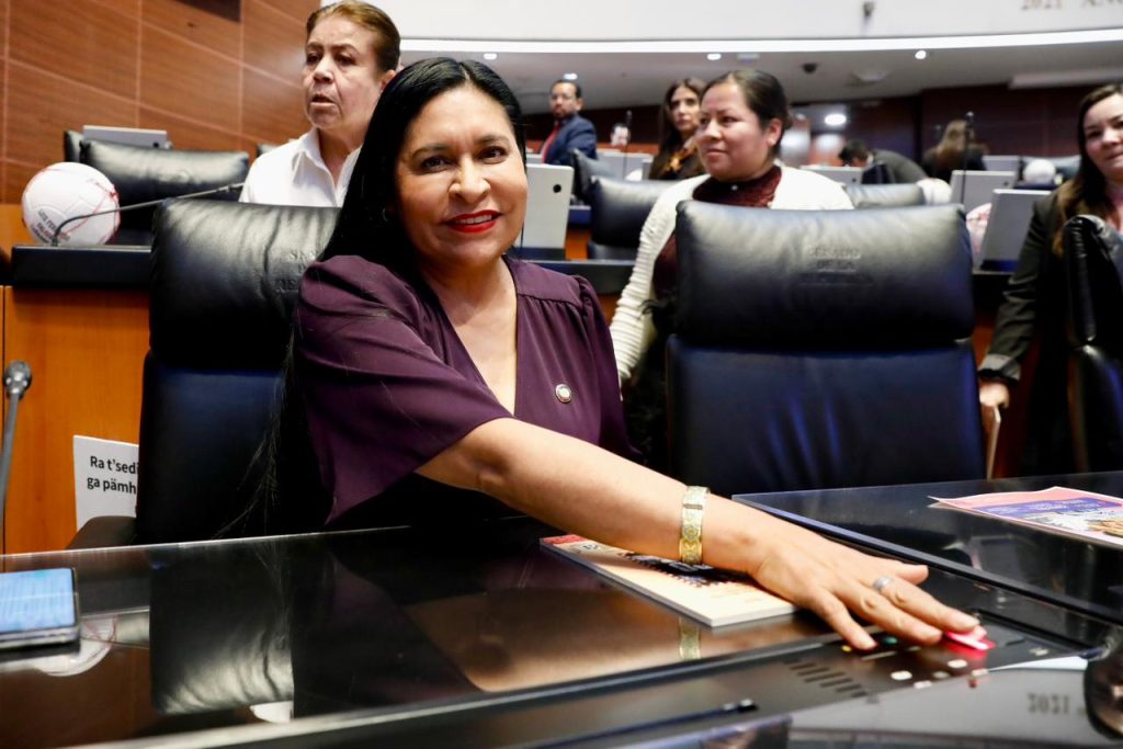 La senadora en una sesión del Congreso, sentada en una silla de cuero, con varias personas en el fondo, en un ambiente formal y de trabajo legislativo.