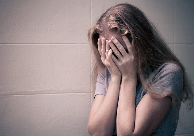 Mujer con cabello rubio y camiseta gris cubriéndose la cara con las manos, mostrando angustia o tristeza, en un fondo de pared beige.