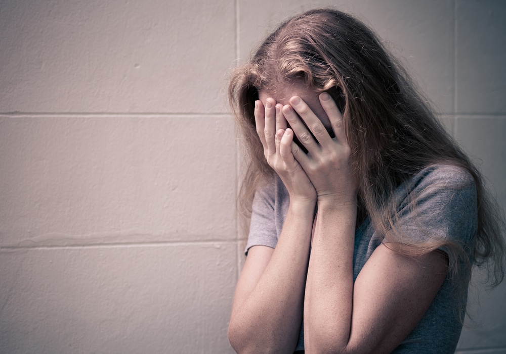 Mujer con cabello rubio y camiseta gris cubriéndose la cara con las manos, mostrando angustia o tristeza, en un fondo de pared beige.
