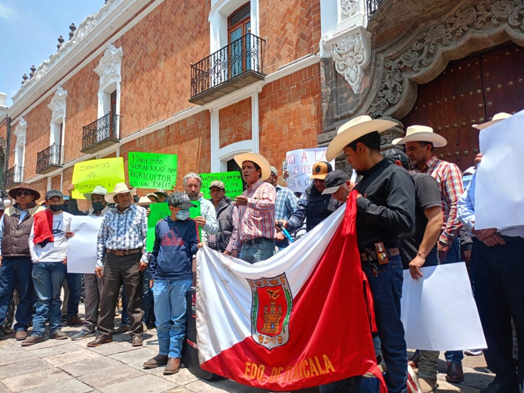 Protesta en Tlaxcala con personas sosteniendo pancartas y banderas, incluyendo la bandera de Tlaxcala, frente a un edificio histórico, en una manifestación por derechos o causas sociales.