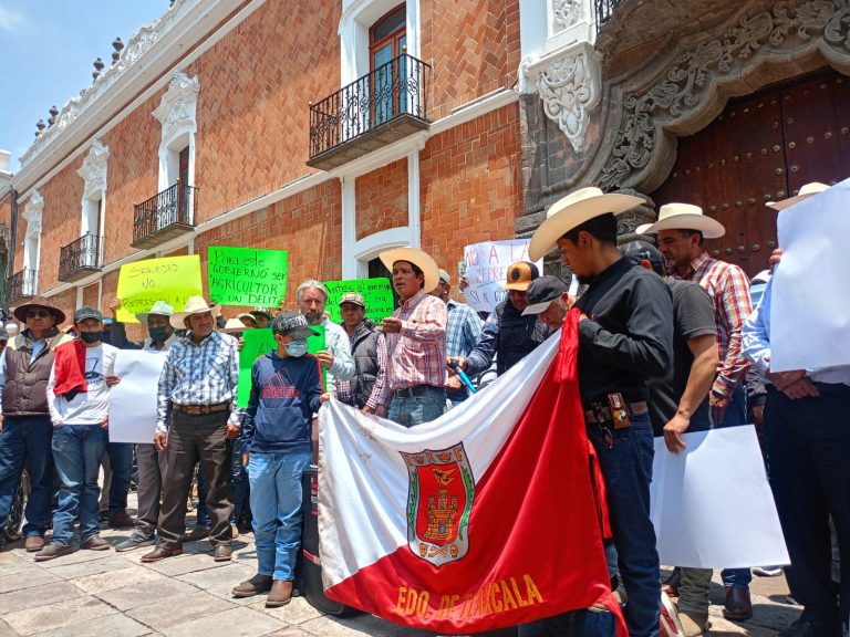 Protesta en Tlaxcala con personas sosteniendo pancartas y banderas, incluyendo la bandera de Tlaxcala, frente a un edificio histórico, en una manifestación por derechos o causas sociales.