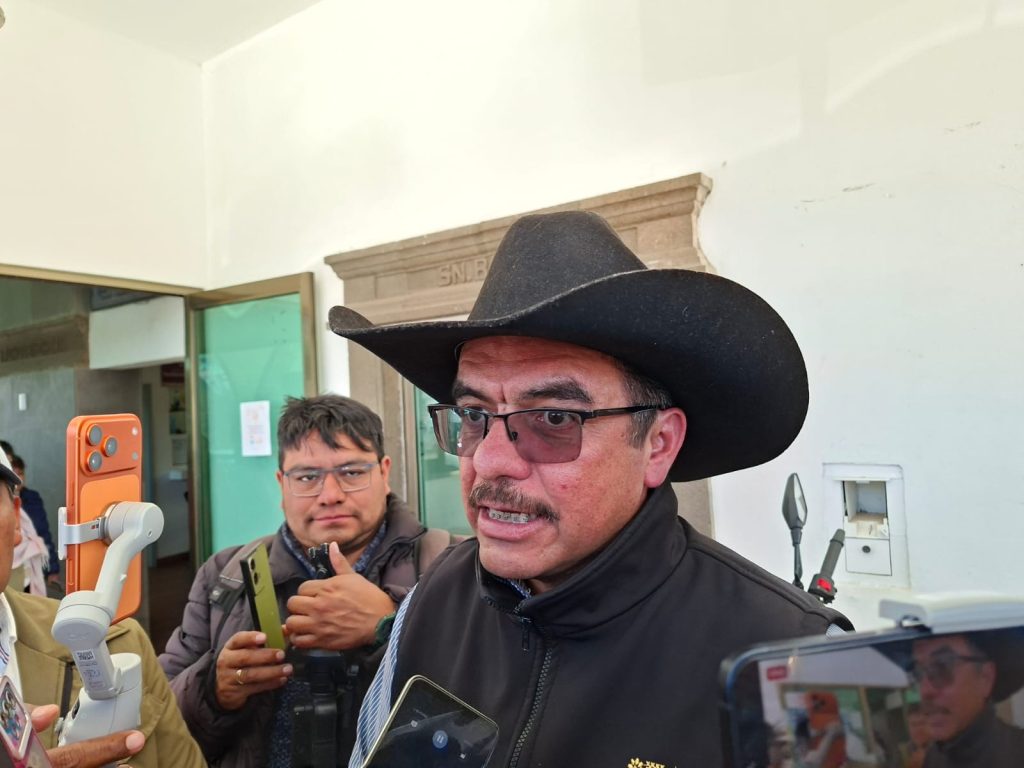 Hombre con sombrero negro y gafas, hablando con medios de comunicación en un entorno interior.