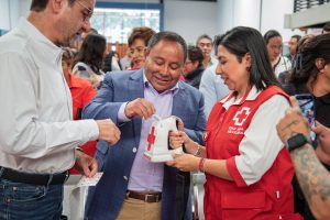Personal de la Cruz Roja entregando un desfibrilador a un hombre en un evento público, con varias personas observando en el fondo.