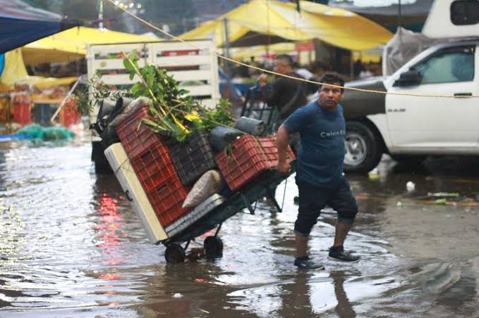 Personas trabajando en un mercado bajo lluvia intensa, con calles inundadas y puestos de venta cubiertos con toldos amarillos, en un entorno urbano durante un día lluvioso en Tlaxcala, México.