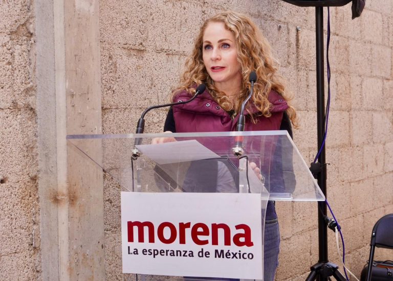 Una mujer con cabello rizado y rubio hablando en un podio con un cartel de Morena, partido político en México, durante un evento público en un entorno exterior con paredes de piedra.