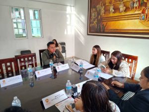 Reunión formal en una sala con cinco personas, incluyendo a un joven en el centro, en una discusión o entrevista, con agua y documentos sobre la mesa.