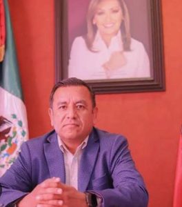 Hombre sentado en una oficina con bandera de México y retrato de mujer en la pared, en un entorno formal y profesional.