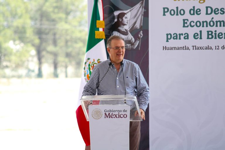 Hombre en podio durante evento oficial en Tlaxcala con bandera de México y cartel de fondo.