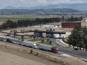Vista de una autopista con camiones y vehículos, rodeada de áreas verdes y un edificio moderno, con montañas al fondo en un día despejado.