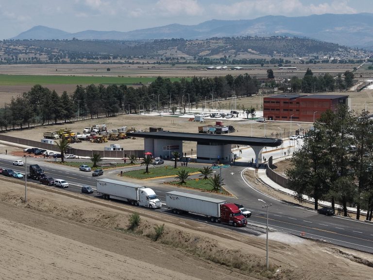 Vista de una autopista con camiones y vehículos, rodeada de áreas verdes y un edificio moderno, con montañas al fondo en un día despejado.