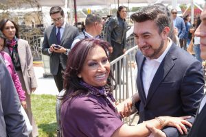 Amplia sonrisa de una mujer y un hombre en un apretón de manos en un evento público, rodeados de varias personas en el fondo, en un ambiente al aire libre.