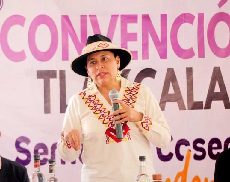 Mujer indígena con sombrero tradicional y vestimenta colorida hablando en una conferencia en Tlaxcala, México.