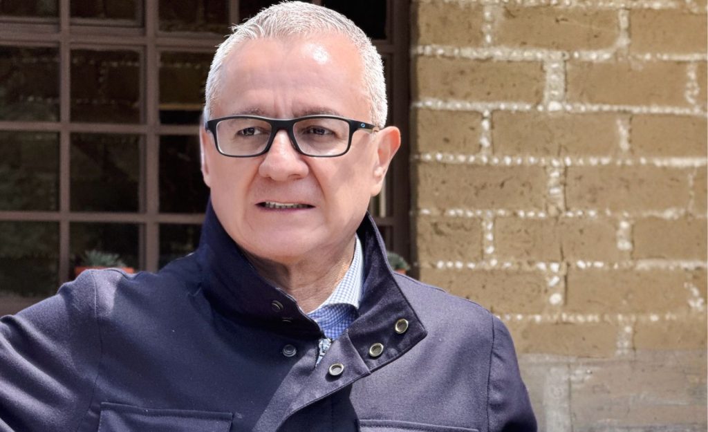 Hombre mayor con gafas y cabello corto gris, vistiendo una chaqueta azul oscuro, de pie frente a una pared de ladrillos, en un entorno urbano.