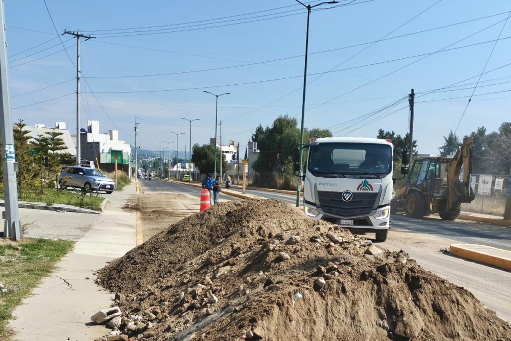Un camión y maquinaria de construcción trabajan en una calle de Tlaxcala, México, con una pila de tierra y señalización vial, en medio de un proyecto de mejora urbana en un día soleado.